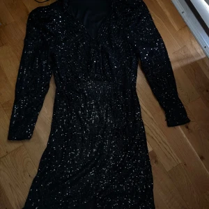 Svart paljettklänning med långa ärmar - Säljer en svart klänning täckt av glittriga paljetter. Klänningen har långa ärmar och markerad midja, perfekt för att sticka ut på festen. Materialet är glansigt och ger en lyxig känsla.