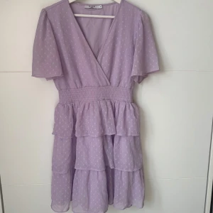 Ljuslila volangklänning från NA-KD - Supersöt ljuslila klänning från NA-KD med volanger och omlottliv. Klänningen har korta ärmar, smock i midjan och prickigt mönster. Perfekt för vår och sommar! Använd en gång!💖💖