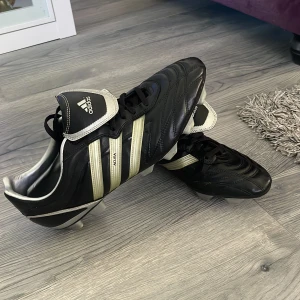 Adidas Copa Mundial fotbollsskor - Klassiska svarta Adidas Copa Mundial fotbollsskor med vita och silvriga detaljer. Skorna har snörning, rund tå och platt sula. Perfekta för dig som vill ha en tidlös look på planen.