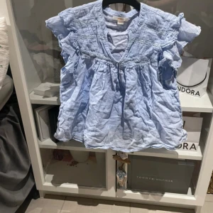 Ljusblå blus med volangdetaljer - Söt ljusblå blus med volangdetaljer på axlarna och smockad överdel. V-ringad hals och lös passform, perfekt för varmare dagar. Tillverkad i ett lätt och luftigt material.