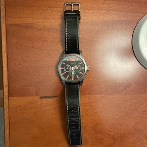 Hugo Boss Klocka  - Stilren klocka från Hugo Boss med svart urtavla och orange detaljer. Klockan har svart läderarmband med vit söm och rostfri boett. Urtavlan visar både datum och veckodag. Perfekt klocka för dig som gillar sportig design. Klockan är även vattentålig. 