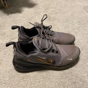 Grå Nike Air Max 270 sneakers - Säljer ett par grå Nike Air Max 270 sneakers med svarta och orange detaljer. Skorna har snörning, rund tå och en synlig luftsula i hälen. Ovandelen är i syntetmaterial med ventilerande mesh. Perfekta för dig som gillar sportig stil.