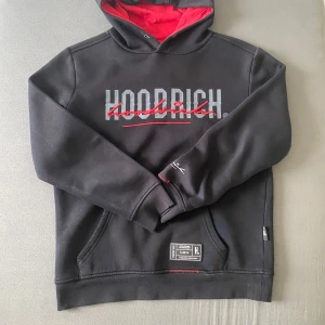 Svart hoodie från Hoodrich med röd luva insidan - Svart hoodie från Hoodrich med stor logga framtill och bak, samt röd insida på huvan. Har en ficka och detaljer i rött och vitt.