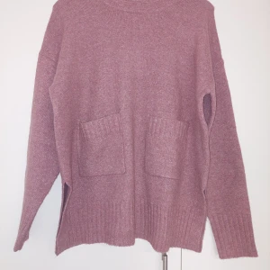 Rosa stickad tröja från KappAhl - Mysig rosa stickad tröja från KappAhl med två stora fickor framtill. Tröjan har lång ärm, rund halsringning och ribbade muddar vid ärmslut och nederkant. Perfekt för kyliga dagar.