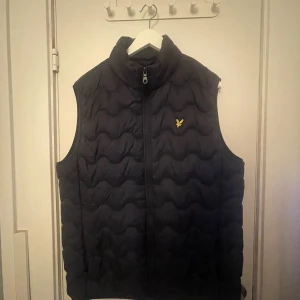 Svart quiltad dunväst från Lyle & Scott - Säljer en svart quiltad dunväst från Lyle & Scott med dragkedja framtill och gul logga på bröstet. Västen har hög krage och ett vågigt quiltat mönster som ger en modern look. Perfekt för lager-på-lager under kyliga dagar.