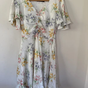 Vit blommig klänning med volangärmar - Söt vit klänning med blommigt mönster i gult, lila och rosa. Klänningen har v-ringning, volangärmar och en lätt rynkad detalj framtill. Perfekt för sommarens alla tillfällen.