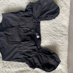 Svart croppad blus med puffärmar - Säljer en svart croppad blus från Divided med korta puffärmar och smockad rygg. Toppen har en söt och trendig design som passar perfekt till jeans eller kjol. Endast använd en gång och är i storlek S. 