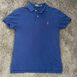 Blå piké från Polo Ralph Lauren - Klassisk blå pikétröja från Polo Ralph Lauren med röd broderad logga på bröstet. Slim fit-modell med korta ärmar, krage och knappar framtill. Perfekt för en stilren och sportig look. Hör av vid funderingar!