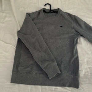 Grå sweatshirt från J.lindeberg - En klassisk grå sweatshirt från J.lindeberg med rund halsringning och diskret logga på bröstet. Tröjan har långa ärmar och ribbade muddar vid ärmslut och nederkant. Perfekt för en avslappnad stil.