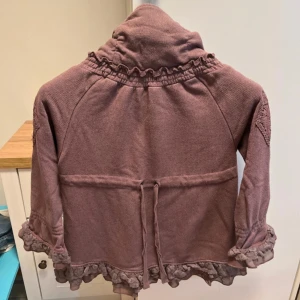 Ljuslila kofta med volang från Cream - Superfin ljuslila kofta från Cream med volangdetaljer längs ärmslut och nederkant. Koftan har hög krage, fickor framtill och knytband i midjan. Perfekt för lager-på-lager och ger en romantisk touch till outfiten.