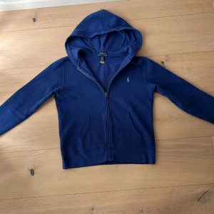 Blå hoodie från Polo Ralph Lauren - Säljer en blå hoodie från Polo Ralph Lauren med dragkedja och huva. Tröjan har två fickor framtill och klassisk broderad logga på bröstet. Perfekt för en avslappnad stil.