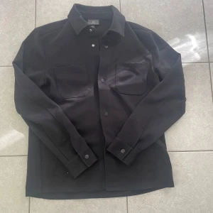 Svart overshirt från H&M - Säljer en svart overshirt från H&M i regular fit. Jackan har två bröstfickor med lock, knäppning framtill och klassisk krage. Perfekt att bära som lager över en t-shirt eller hoodie.