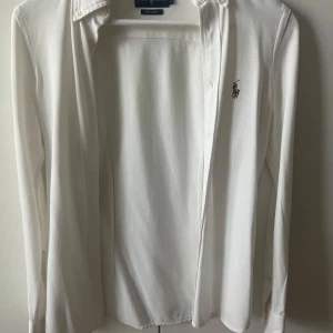 Vit långärmad skjorta från Ralph Lauren - Klassisk vit långärmad skjorta från Ralph Lauren med broderad logga på bröstet. Skjortan är i mjuk bomull och har knappar framtill samt button-down krage. Perfekt för en stilren look.