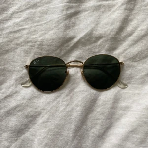 Ray-Ban runda solglasögon - Säljer ett par Ray-Ban solglasögon med runda, mörkgröna glas och tunna guldfärgade metallbågar. Klassisk och tidlös modell med näsbrygga i metall och transparenta nässkydd. Perfekt accessoar för soliga dagar.