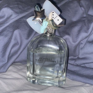 Perfect Eau de Toilette från Marc Jacobs - Säljer en flaska Perfect Eau de Toilette från Marc Jacobs, 100 ml. Flaskan är genomskinlig med unika dekorationer på korken som stjärna, dominobricka och pastellfärgade detaljer. Doften är fräsch och modern, perfekt för dig som gillar stilrena och lekfulla parfymer.