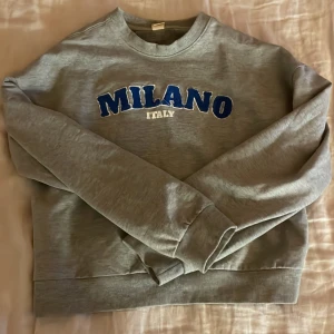 Grå sweatshirt med Milano-tryck från Uniq Young - Säljer en grå sweatshirt från Uniq Young med blått och vitt Milano-tryck på bröstet. Tröjan har rund halsringning och långa ärmar. Perfekt för en avslappnad stil.