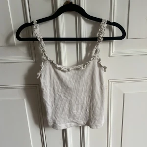 Vit croppat linne med volangdetaljer - Säljer ett söt vit croppat linne med volangdetaljer längs axelbanden och urringningen. Tunt ribbat material vilket gör det lite mer uppklätt. Perfekt till varma dagar!