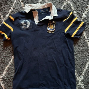 Marinblå pikétröja från Polo Ralph Lauren - Säljer en marinblå pikétröja från Polo Ralph Lauren med klassisk vit krage och gula ränder på ärmarna. Tröjan har broderad logga och klubbmärke på bröstet samt nummerdetalj på axeln. Perfekt för en vintage stil!