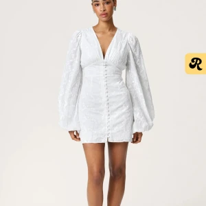 Vit kortklänning med puffärmar och knappar - Superfin vit kortklänning med långa puffärmar och dekorativa knappar framtill. Klänningen har diskret blommigt mönster i tyget och markerad midja för en feminin siluett. Perfekt för sommarens tillfällen! 