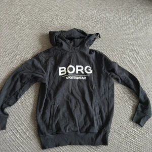 Svart hoodie från Björn Borg - Säljer en svart hoodie från Björn Borg med stor vit logga 'BORG SPORTSWEAR' på bröstet. Tröjan har huva och långa ärmar samt ribbade muddar vid ärmslut och nederkant. Perfekt för en sportig och avslappnad stil.