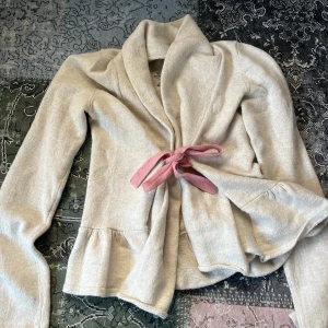 Beige stickad cardigan jacka från Odd Molly - Mysig beige stickad cardigan jacka från Odd Molly med knytband i rosa framtill och broderad logga på ryggen. Jackan har lång ärm och volangdetalj nedtill. Perfekt för lager-på-lager-stil. Storlek 0 som motsvarar xs. Pris går alltid att diskutera