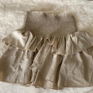 Beige volangkjol från Neo Noir - Supersöt beige kjol från Neo Noir med bred smock i midjan och dubbla volanglager. Kjolen har en kort modell och är perfekt för varma dagar. Lätt och luftig känsla med snyggt fall.