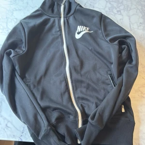 Svart zip-jacka från Nike - Svart jacka från Nike med vit dragkedja och logga på bröstet. Jackan har ribbade muddar, hög krage och två fickor med dragkedja. Perfekt för en sportig look och enkel att matcha med det mesta. Köpt på Nike 799 kr mitt pris 250 kr mycket bra pris🤩
