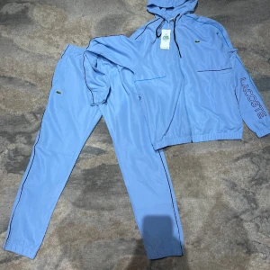 Baby blå Lacoste tracksuit  - Säljer en ljusblå tracksuit från Lacoste med matchande byxor och jacka med huva storlek M. Jackan har dragkedja, snörning i huvan, Lacoste-logga på bröstet och stor krokodil på ryggen. Byxorna har resår i midjan och liten logga på benet. Org pris 2599, mitt pris 1800 priset kan också sänkas för mer bilder skriv gärna