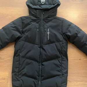 Svart dunjacka från Sail Racing - Säljer en svart dunjacka från Sail Racing med huva och dragkedja framtill. Jackan har två sidofickor och en bröstficka med dragkedja samt logga på bröstet. Perfekt för kalla dagar och riktigt snygg stil.