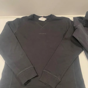 En Svart sweatshirt och en blå hoodie från Les Deux - Två svarta tröjor från Les Deux – en klassisk sweatshirt med diskret broderad logga på bröstet och en hoodie med broderad detalj. Båda har långa ärmar och ribbade muddar. Perfekta för en stilren och avslappnad look.