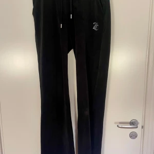 Svarta mjukisbyxor från Juicy Couture - Svarta mjukisbyxor från Juicy Couture med raka ben och dragsko i midjan. Byxorna har Juicy-logga i strass på baksidan och en liten logga framtill. Perfekta för en avslappnad och trendig look. Lite små slit här å där men inget man tänker på jätte mycket 