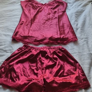 Rosa satinlinne med spetsdetaljer - Säljer VinRöt linne i satin med spetsdetaljer vid halsringning och nederkant. Linnet har en feminin v-ringning och ärmlös design, perfekt för en mjuk och lyxig känsla. Shortsen följer också med spets på nederdelen 