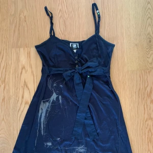 Mörkblå klänning med knytband från G-Star RAW - Svart klänning från G-Star RAW med smala axelband, knappar framtill och knytband i midjan. Klänningen har ett tryckt motiv framtill och är tillverkad i mjukt material. Perfekt för en stilren och avslappnad look.