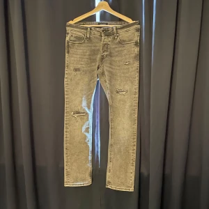 Grå jeans Jack&Jones med slitningar - Säljer ett par grå jeans med slitna detaljer på framsidan. Byxorna har klassisk femficksdesign och raka ben. Perfekta för en avslappnad och trendig look.