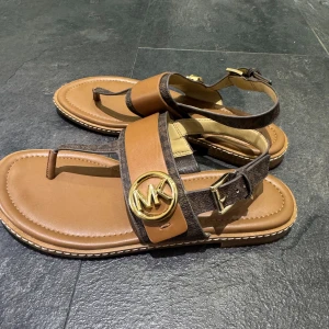 Bruna sandaler från Michael Kors med gulddetalj - Snygga sandaler från Michael Kors i brunt skinn och syntet med bred rem och stort MK-emblem i guld framtill. Justerbart spänne vid ankeln och bekväm platt sula. Perfekta för varma dagar och enkel att matcha med olika outfits.