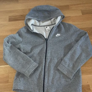 Grå hoodiejacka från Nike - Grå zip-hoodie från Nike med vit broderad logga på bröstet. Jackan har huva, dragkedja framtill och två fickor. Mjuk insida och klassisk sportig look, perfekt för vardagsbruk.