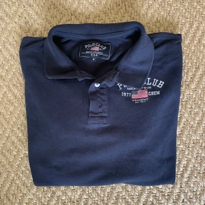 Mörkblå pikétröja från Polo Club - Säljer en klassisk mörkblå pikétröja från Polo Club Saint Vincent med broderad logga och amerikansk flagga på bröstet. Tröjan har krage, knappar framtill och korta ärmar. Perfekt för en stilren och sportig look.