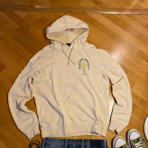 Beige hoodie från Les Deux med Sunrise Society-tryck - Säljer en beige hoodie från Les Deux med grönt och gult Sunrise Society-tryck både fram och bak. Tröjan har huva med dragsko och ribbade muddar. Perfekt för en avslappnad stil.
