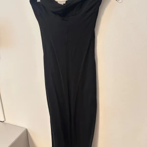 Svart långklänning med draperad halsringning - Elegant svart långklänning med draperad halsringning och smal passform. Klänningen är ärmlös och har en stilren, tidlös design som passar perfekt till festliga tillfällen.