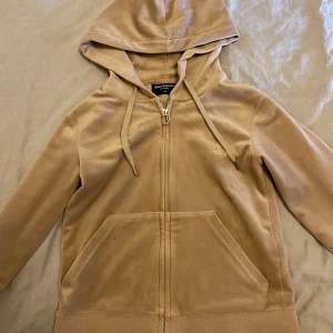 Beige velour hoodie från Juicy Couture - Säljer en beige hoodie i mjuk velour från Juicy Couture. Jackan har dragkedja framtill, huva med dragsko och två fickor. Klassisk Juicy Couture-logga på dragkedjan och liten broderad detalj på bröstet.