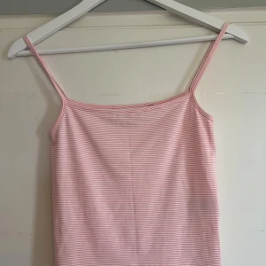 Rosa och vit randig linnetopp - Säljer en söt rosa och vit randig linnetopp med tunna axelband. Perfekt för varma dagar och enkel att matcha med jeans eller kjol. Toppens material känns mjukt och bekvämt mot huden.