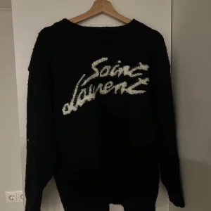 Svart stickad tröja från Saint Laurent - Säljer en svart stickad tröja från Saint Laurent med vit logotyp framtill. Tröjan har rund halsringning och långa ärmar. Perfekt för dig som vill ha en statement piece i garderoben.