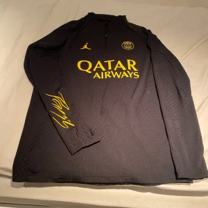PSG träningströja - Säljer en svart långärmad PSG fotbollströja från Jordan med gul logga och detaljer. Tröjan har halv dragkedja, Paris Saint-Germain-emblem på bröstet och Qatar Airways som sponsortryck. Slim fit-modell, perfekt för fotboll eller streetwear. Detta är de tröjor som används av proffsen vilket höjer priset. Skillnaden mellan proffströjorna och vanliga tröjor är materialet, mönstret och passformen