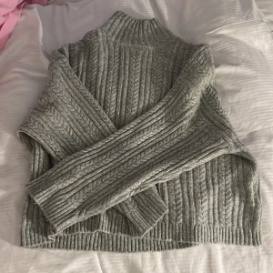 Grå stickad polotröja från ICHI - Säljer en grå stickad polotröja från ICHI. Tröjan har ett klassiskt kabelstickat mönster, hög krage och långa ärmar. Perfekt för kyliga dagar och enkel att matcha med olika outfits.