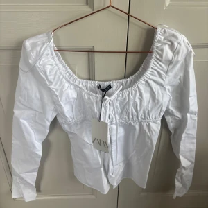 Vit långärmad blus från Zara - Säljer en vit långärmad blus från Zara med bred, rynkad urringning och figurnära passform över bysten. Blusen har en romantisk känsla och är tillverkad i ett mjukt bomullstyg. Perfekt att styla med jeans eller kjol.