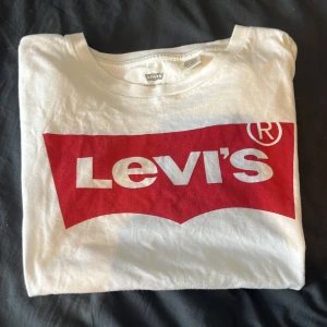 Vit t-shirt med röd Levi's-logga - Vit t-shirt från Levi's med den klassiska röda loggan tryckt framtill. T-shirten har rund halsringning och korta ärmar. Perfekt för en avslappnad och stilren look.