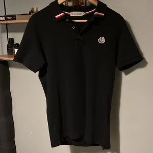 Svart pikétröja från Moncler - Snygg svart pikétröja från Moncler med klassisk krage och knäppning framtill. Tröjan har Moncler-logga på bröstet och kragen har röda och vita detaljer. Perfekt för en stilren look.