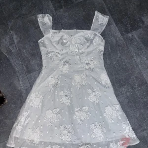 Vit blommig klänning från Cider - Supersöt vit klänning från Cider med blommigt broderi och volangärmar. Klänningen har knytning framtill och är tillverkad i polyester. Perfekt som sommarklänning eller till student 😍