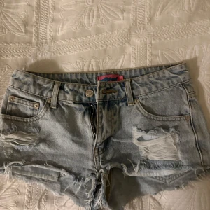 Ljusblå jeansshorts med slitningar - Säljer ett par ljusblå jeansshorts med råa kanter och slitna detaljer framtill. Klassisk femficksmodell med knapp och dragkedja. Perfekta för varma dagar och en avslappnad stil. Använda en gång då de var för små