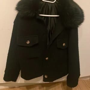 Svart kappa med päls-krage  - Säljer en svart kappa med stor krage i päls och dekorativa knappar framtill. Jackan har två fickor med lock och är helfodrad. Perfekt för dig som vill ha en klassisk och snygg look.